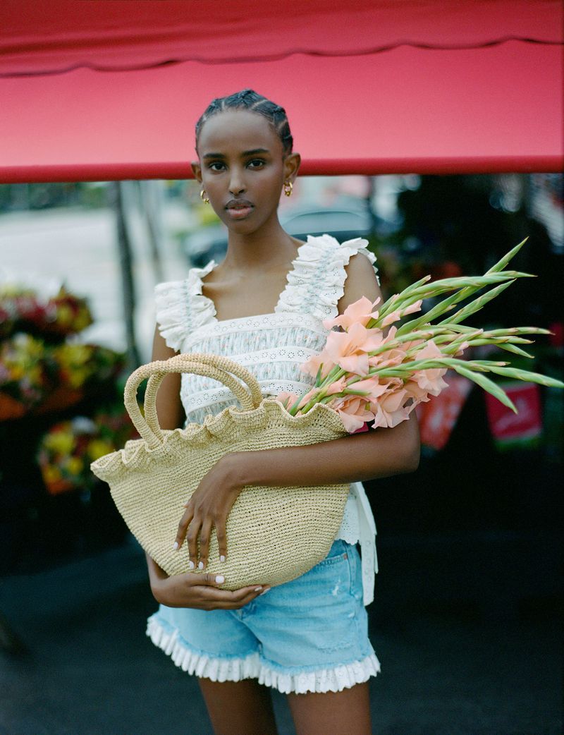 A woman poses holding a straw bag filled with flowers in a fashion shot; she wears a white top and blue jean shorts with ruffles.