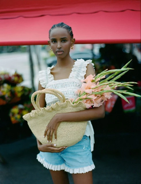 A woman poses holding a straw bag filled with flowers in a fashion shot; she wears a white top and blue jean shorts with ruffles.