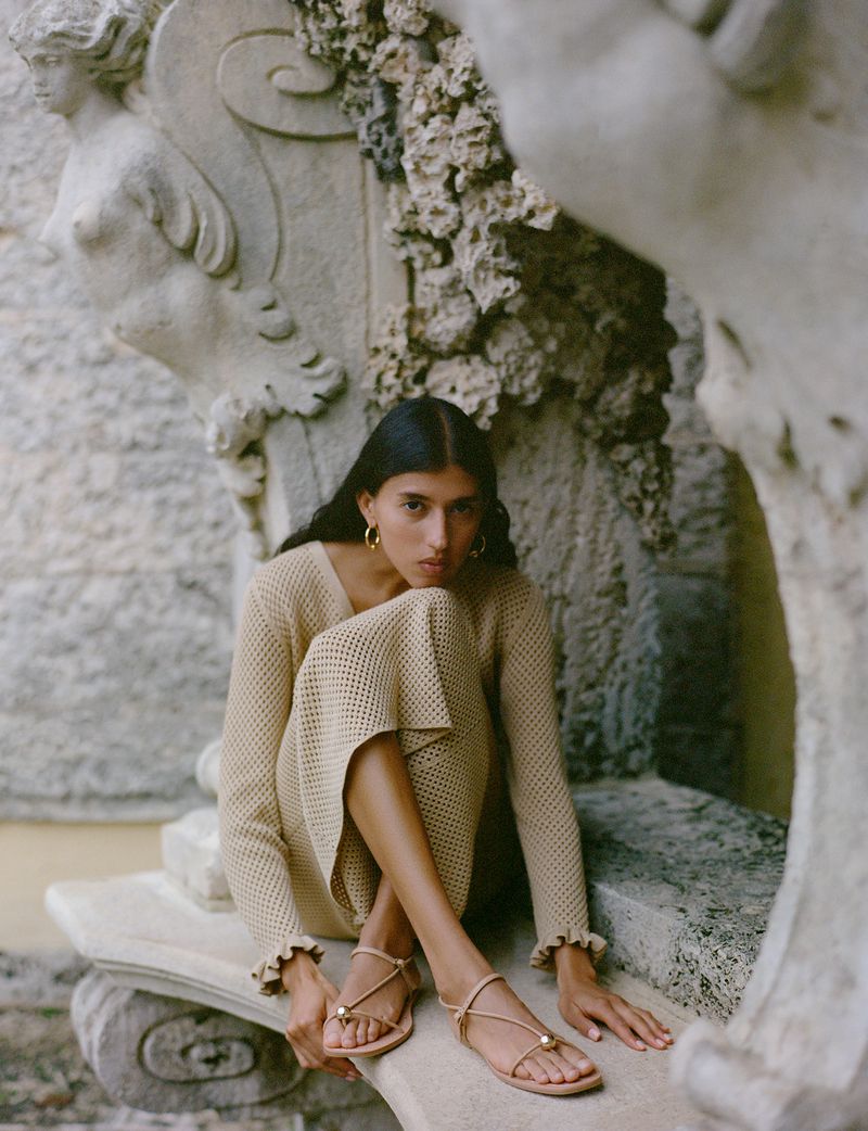 Woman in beige knitted dress and strappy sandals poses near weathered stone fountain in a sunlit outdoor setting, creating a serene portrait.
