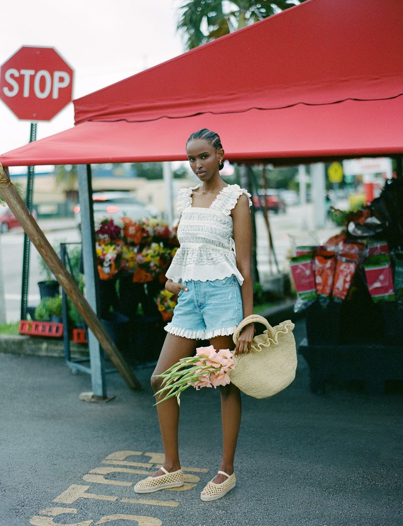 A dark-skinned woman stands outdoors holding flowers and a woven bag, wearing a white top and blue shorts. A red stop sign is visible.