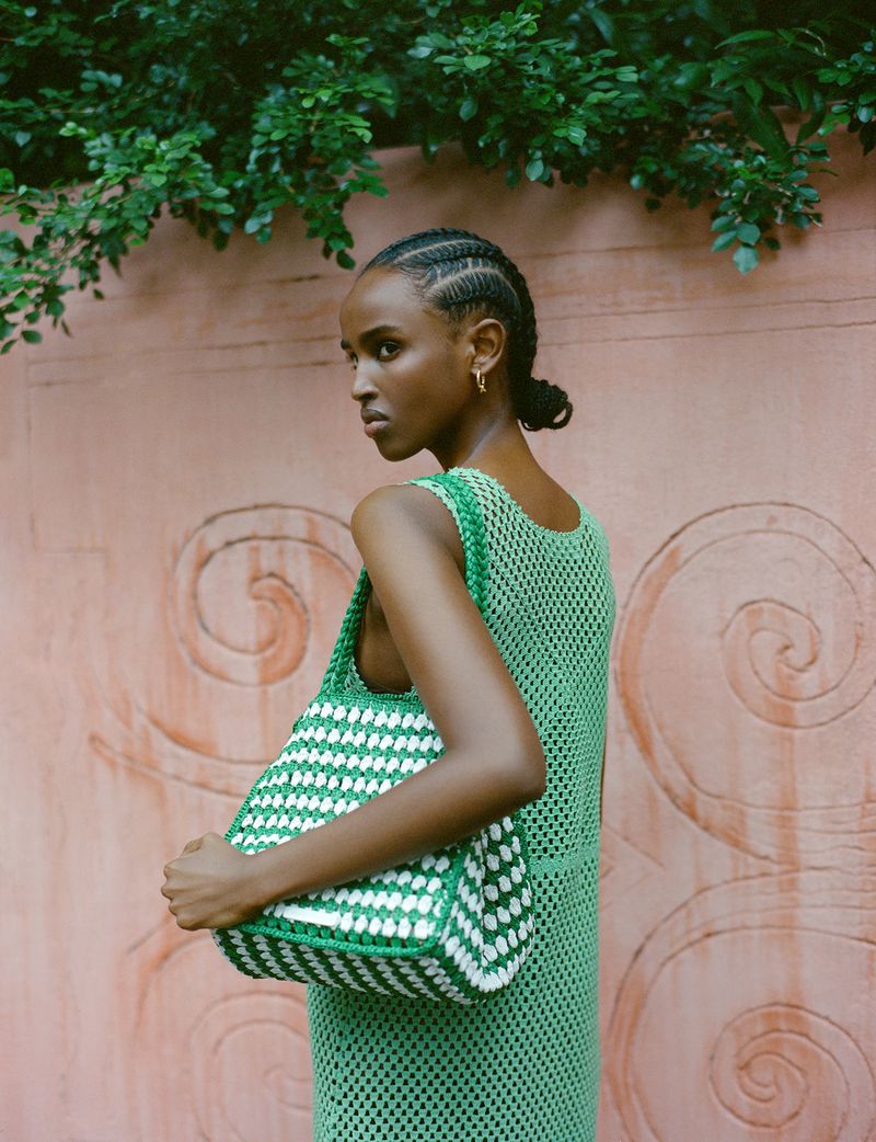 Woman in green crochet dress holds a green and white woven tote bag, posing outdoors against a textured pink wall with green foliage.