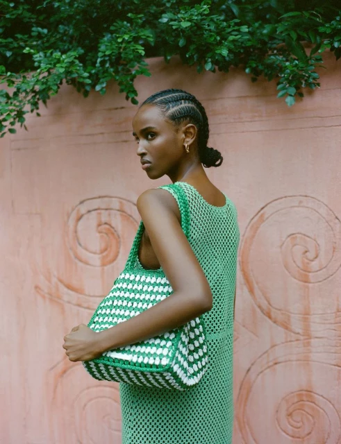 Woman in green crochet dress holds a green and white woven tote bag, posing outdoors against a textured pink wall with green foliage.
