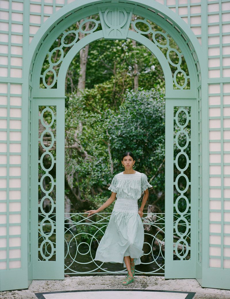 Adult woman in a light green dress posing in front of a light green, arched trellis with greenery behind her in a medium shot.