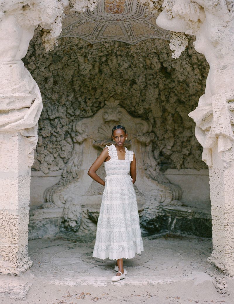 A light-skinned woman models a white cotton dress against a carved stone background with statues and fountain, creating a vintage fashion look.
