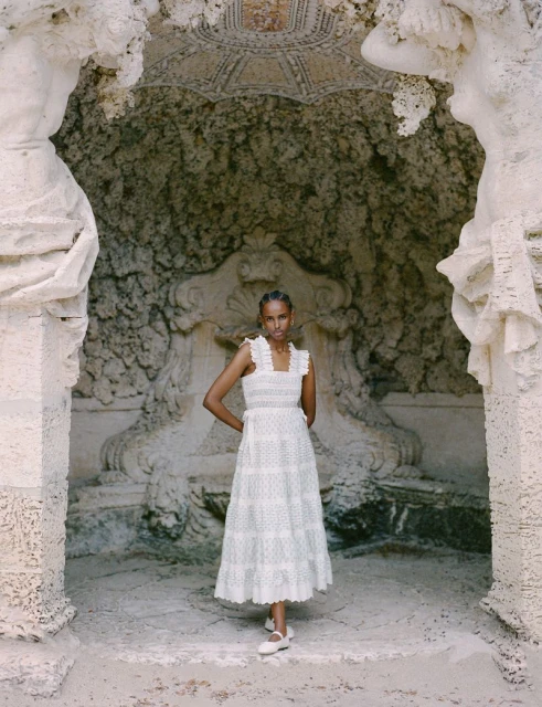 A light-skinned woman models a white cotton dress against a carved stone background with statues and fountain, creating a vintage fashion look.