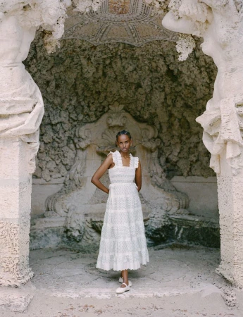 A light-skinned woman models a white cotton dress against a carved stone background with statues and fountain, creating a vintage fashion look.