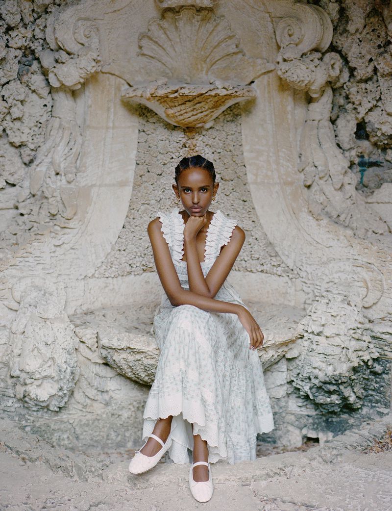 Elegant woman in a floral dress sits gracefully on a stone structure with an ornamental wall in the background, looking straight at the camera.