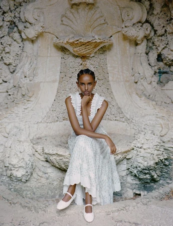 Elegant woman in a floral dress sits gracefully on a stone structure with an ornamental wall in the background, looking straight at the camera.