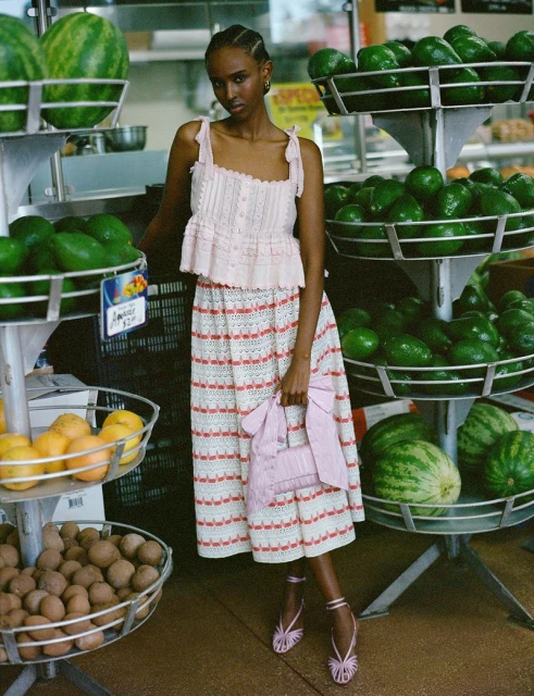 A fashionable woman stands in a market in a pink outfit, posing near displays of watermelons, avocados, oranges, and other fresh produce.