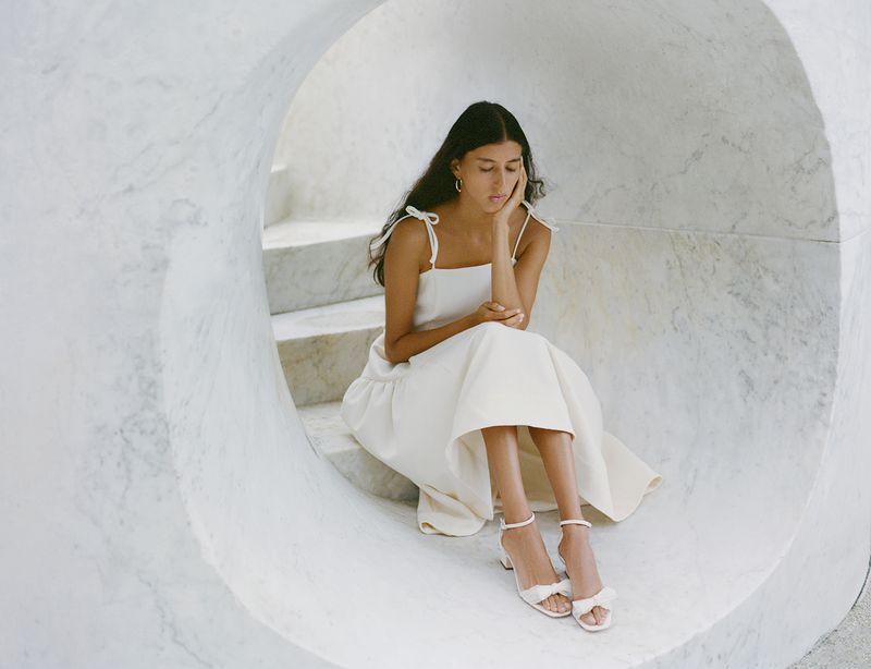 Woman in a cream-colored sundress and matching heels poses in a marble architectural setting with diffused lighting indoors.