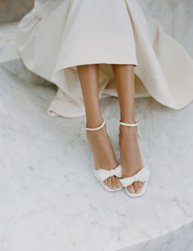 Close-up studio shot of a woman in a white dress and white strappy heeled sandals with bows, sitting on light marble steps.