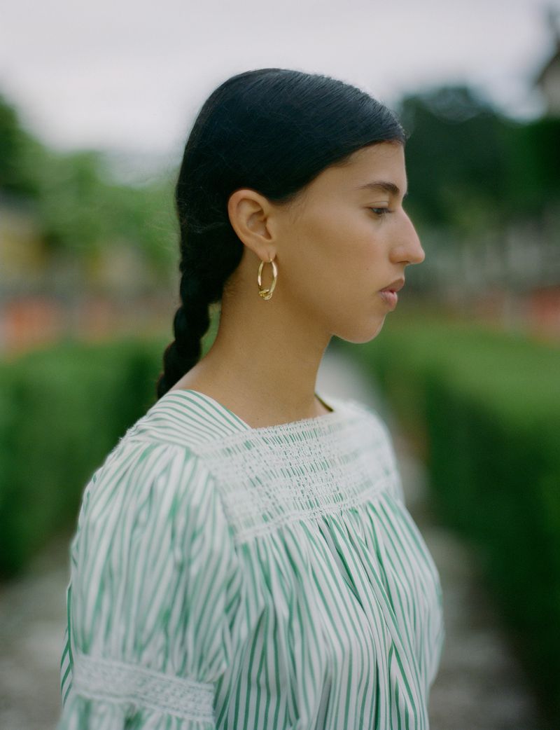 Woman in a vertically striped blouse with gold hoop earrings poses in an outdoor garden setting on an overcast day.