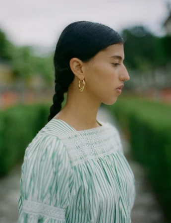 Woman in a vertically striped blouse with gold hoop earrings poses in an outdoor garden setting on an overcast day.