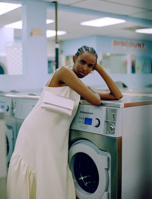 A woman with braided hair wearing a dress stands next to a washing machine in a laundromat, looking directly at the camera.