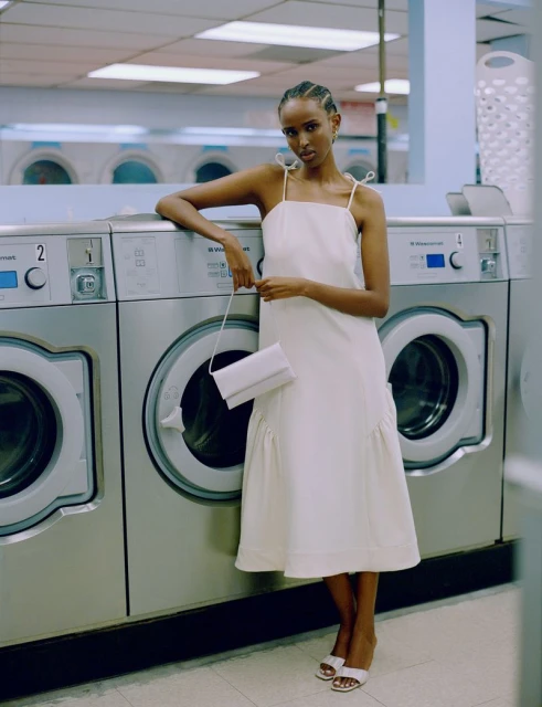 An elegant woman poses in a laundromat. She's wearing a white dress with thin straps, holding a white purse, with washing machines behind her.