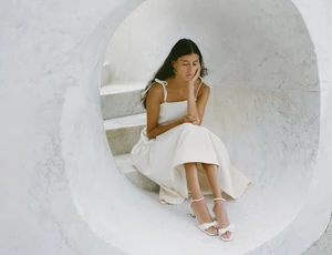 Model seated inside a circular marble tunnel wearing a white tie-shoulder dress and white bow block-heel sandals.
