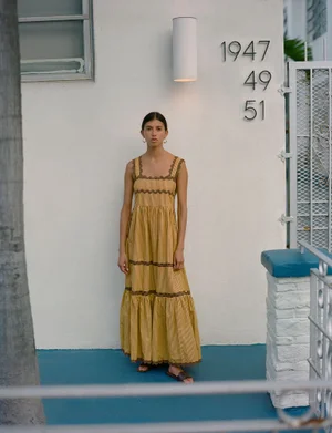 Model wearing a yellow tiered maxi dress and flat sandals standing against a white building exterior.