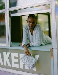 A woman leans out of a storefront window wearing a pale blue knit cardigan and holding a light blue woven clutch bag.
