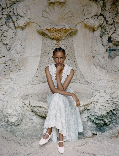 Model Ifrah Qaasim wearing a ruffled white maxi dress and cream ballet flats, seated in an ornate stone grotto niche.
