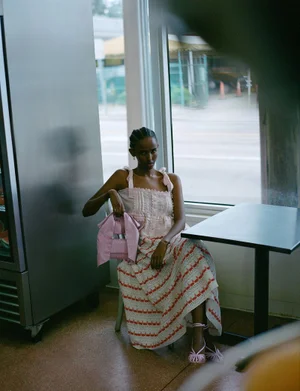 Model seated indoors holding a pink handbag wearing a white dress with red patterned stripes