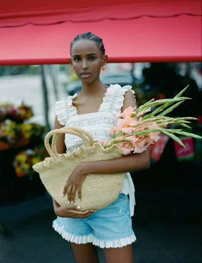 Model holding a woven straw tote filled with peach gladiolus flowers under a red awning for Loeffler Randall SU24.