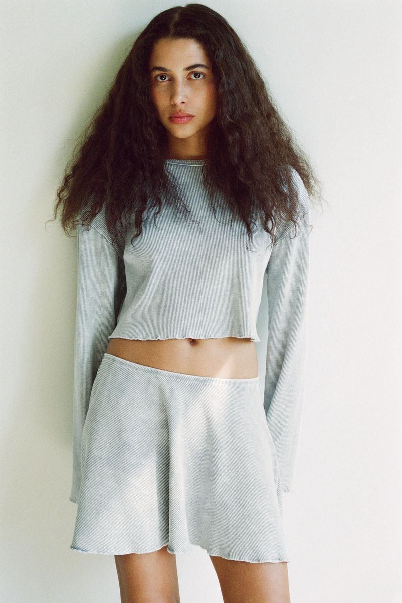 Woman poses in a cropped long sleeve shirt and skirt set indoors. The clothing is a textured light blue. The background is a plain wall.