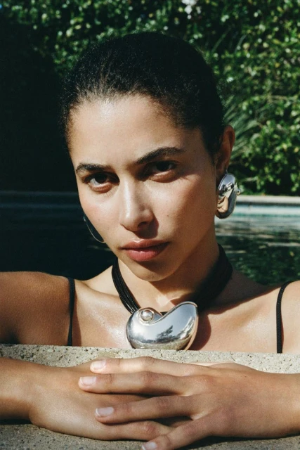Close-up of a woman with dark hair and silver jewellery posing in front of a green, outdoor background on a sunny day.