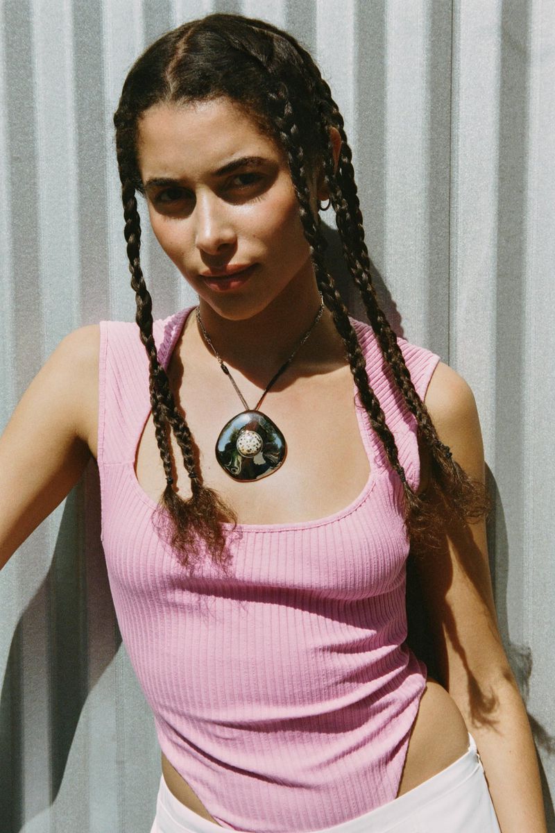 Portrait of a young woman with braids wearing a pink top and necklace, standing in front of a corrugated metal wall, bathed in natural light.