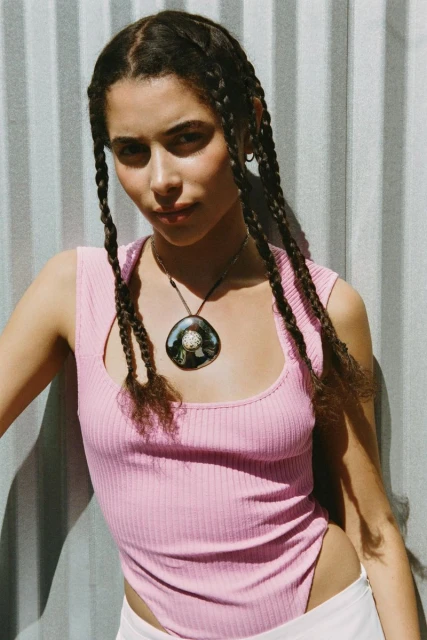 Portrait of a young woman with braids wearing a pink top and necklace, standing in front of a corrugated metal wall, bathed in natural light.