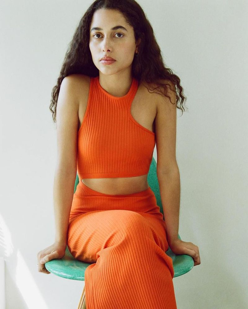 Young adult woman with long dark curly hair sitting on a light blue chair wearing an orange knit skirt and matching crop top.
