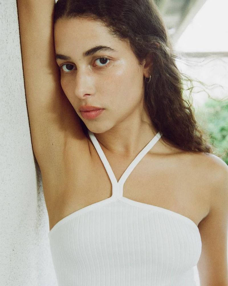 Close-up portrait of a young woman with curly brown hair, wearing a white tank top, posing against a light grey textured wall, neutral expression.