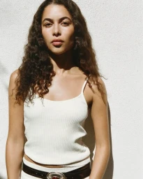 Model Alici Sol posing in a white ribbed camisole and leather belt against a sunlit textured wall.