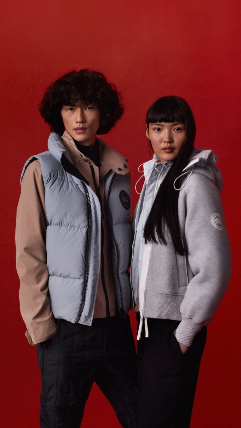 Two Asian individuals, a man and a woman, model winter fashion against a vibrant red backdrop. The man sports a puffer vest.