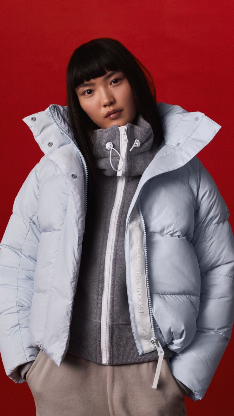 Portrait of a young Asian woman wearing a light-blue puffer jacket, grey sweater, and taupe pants against a red backdrop.