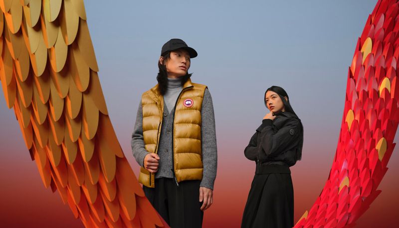 Two models pose against a background of orange and red paper scales, showcasing golden puffer vest and black jacket in a studio setting.