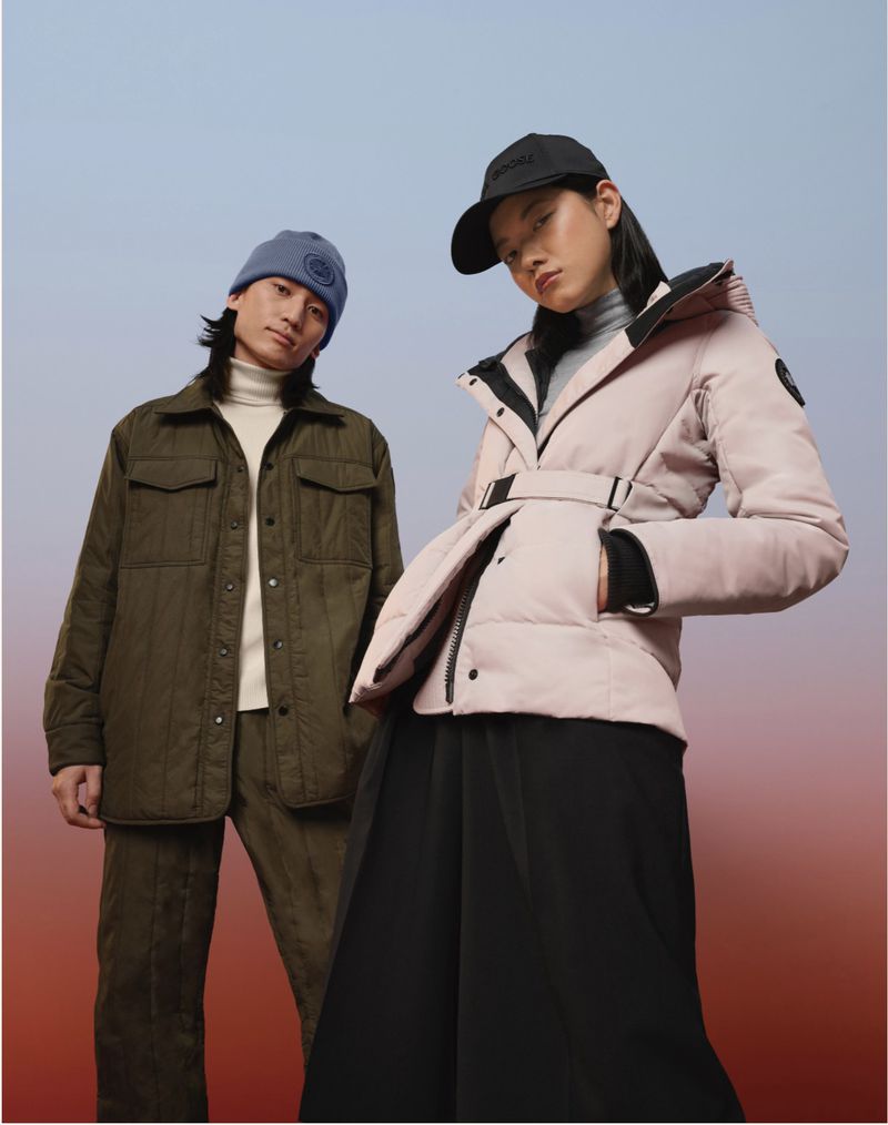 Two fashion models pose in winter apparel against a blended color gradient background. One wears a green jacket and pants, the other a pink coat.