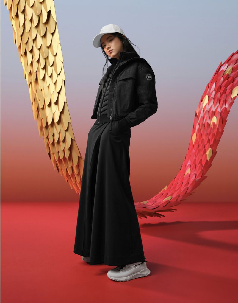 A fashionable young woman in a black jacket, long skirt, cap, and sneakers poses in a studio with a red floor and gold and red decorations.
