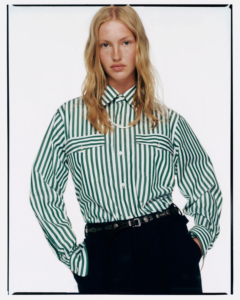 Fashion shot of a fair-skinned woman with blond hair wearing a green and white striped button-down shirt and black pants in a photo studio.