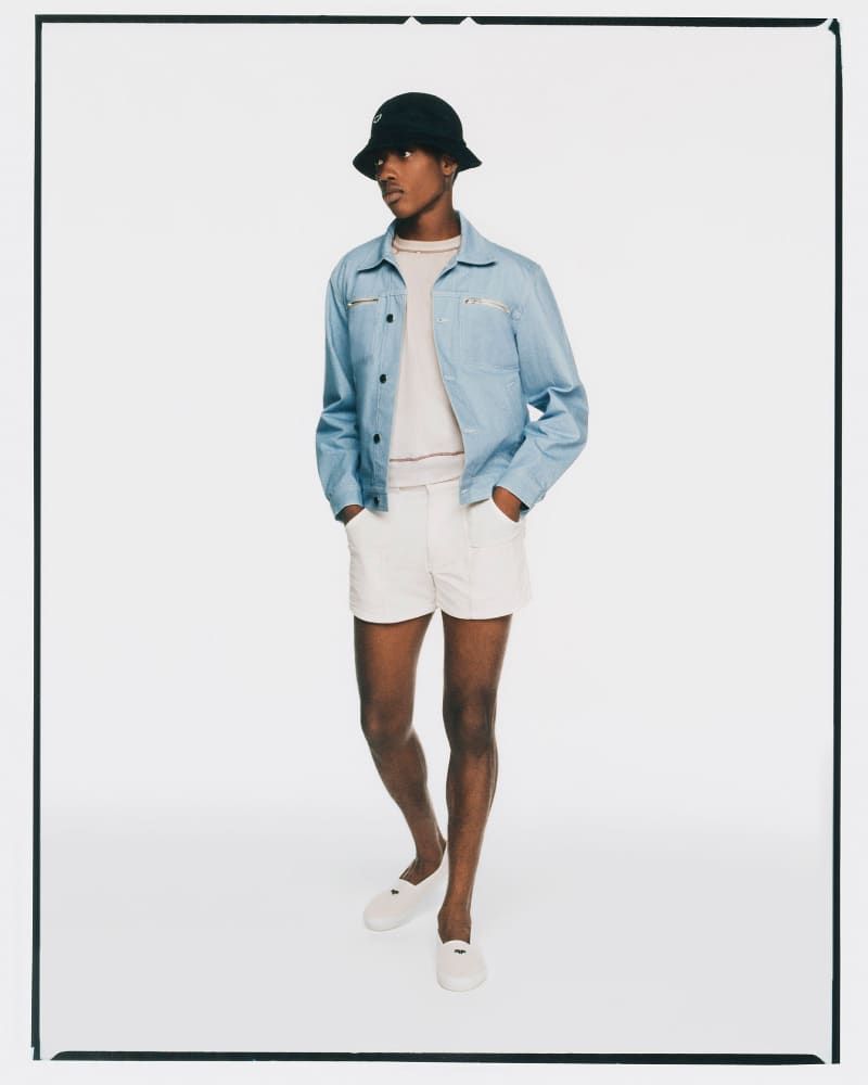 Full-length shot of a young adult male wearing a light blue jacket, white shorts, bucket hat, and loafers, posing in a photo studio.