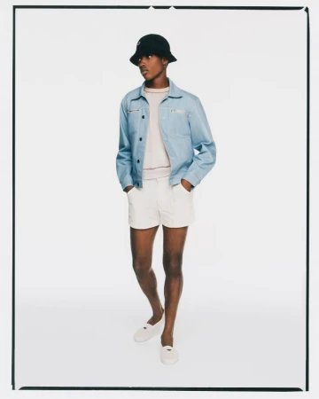 Full-length shot of a young adult male wearing a light blue jacket, white shorts, bucket hat, and loafers, posing in a photo studio.
