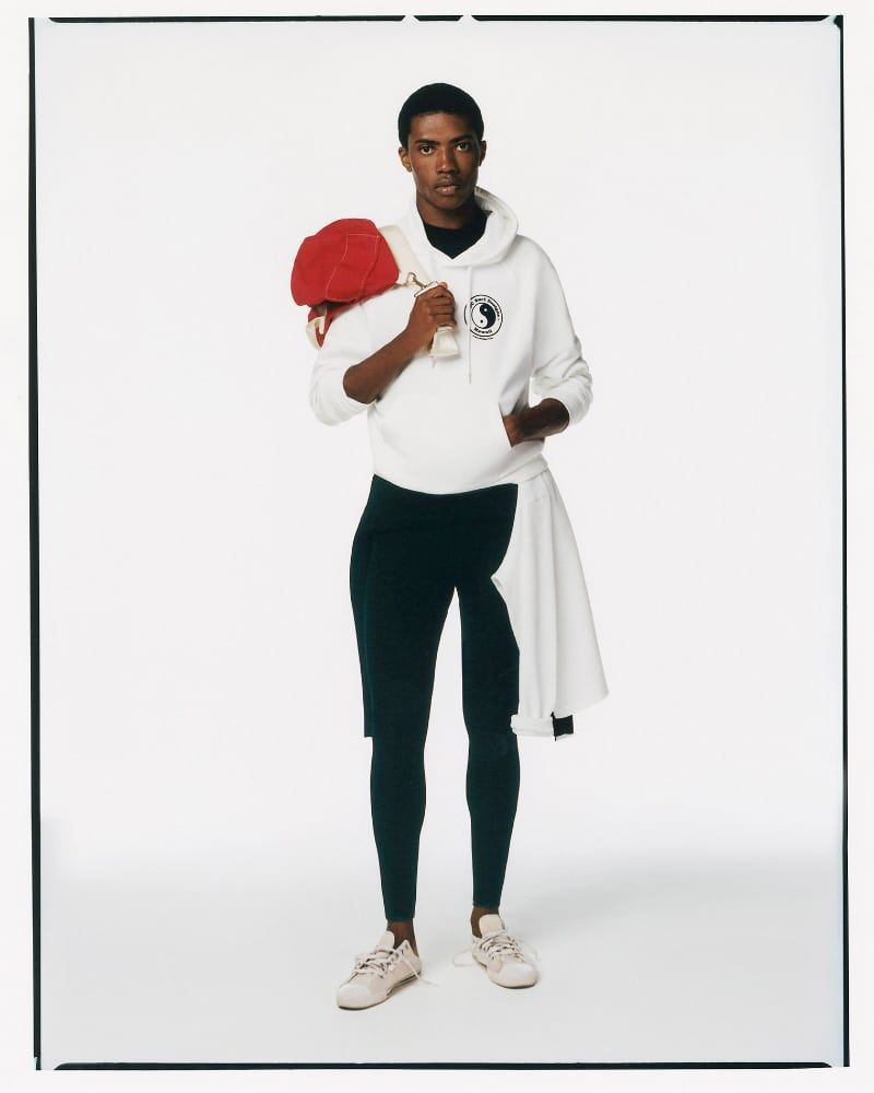 Full shot of an African-American man in a white hoodie and dark leggings, holding a red ball over his shoulder in a white studio.