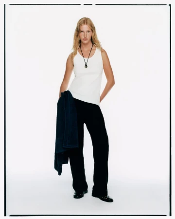 A fair-skinned woman with blonde hair is standing in a white photo studio wearing a white tank top, black pants, and black shoes.