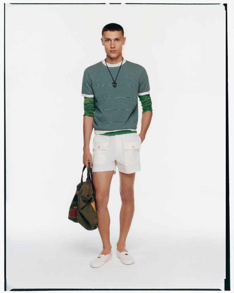 Full-length shot of a male model in a striped green and white shirt, white shorts, white shoes, and holding a duffel bag.