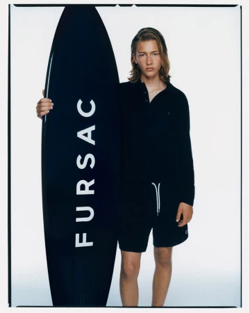 Young man in black polo shirt and shorts stands next to a black surfboard with white text on a white background.