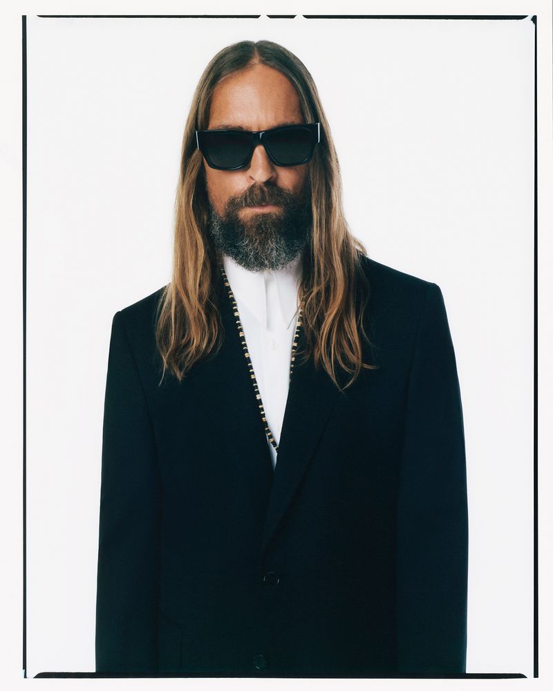 Portrait of a bearded man with long hair wearing a black suit and sunglasses against a bright white studio background. Male fashion.