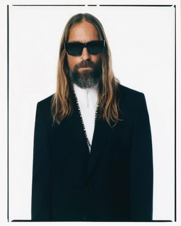 Portrait of a bearded man with long hair wearing a black suit and sunglasses against a bright white studio background. Male fashion.