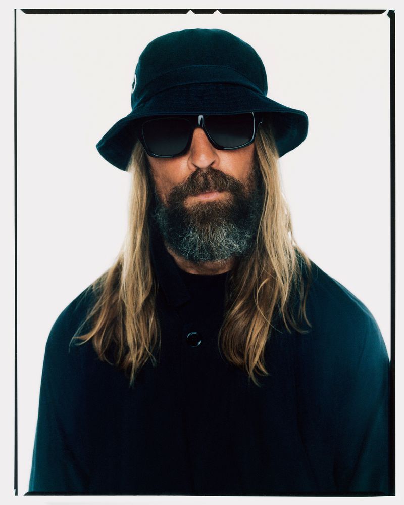 Portrait of a male adult with long hair and a beard, wearing sunglasses and a hat, posing in front of a white backdrop.