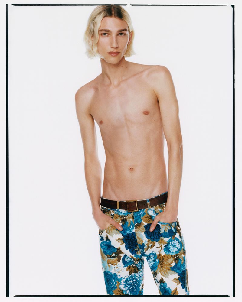 Blonde male fashion model poses shirtless in a studio with floral pants, looking at the camera in a fashion studio environment.