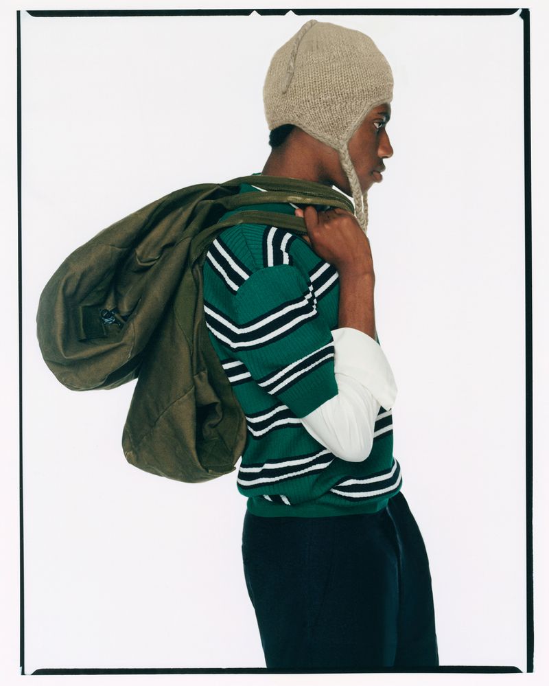Side view of a Black male carrying an olive green duffel bag over his shoulder, wearing a knit hat and striped green sweater, against a white backdrop.