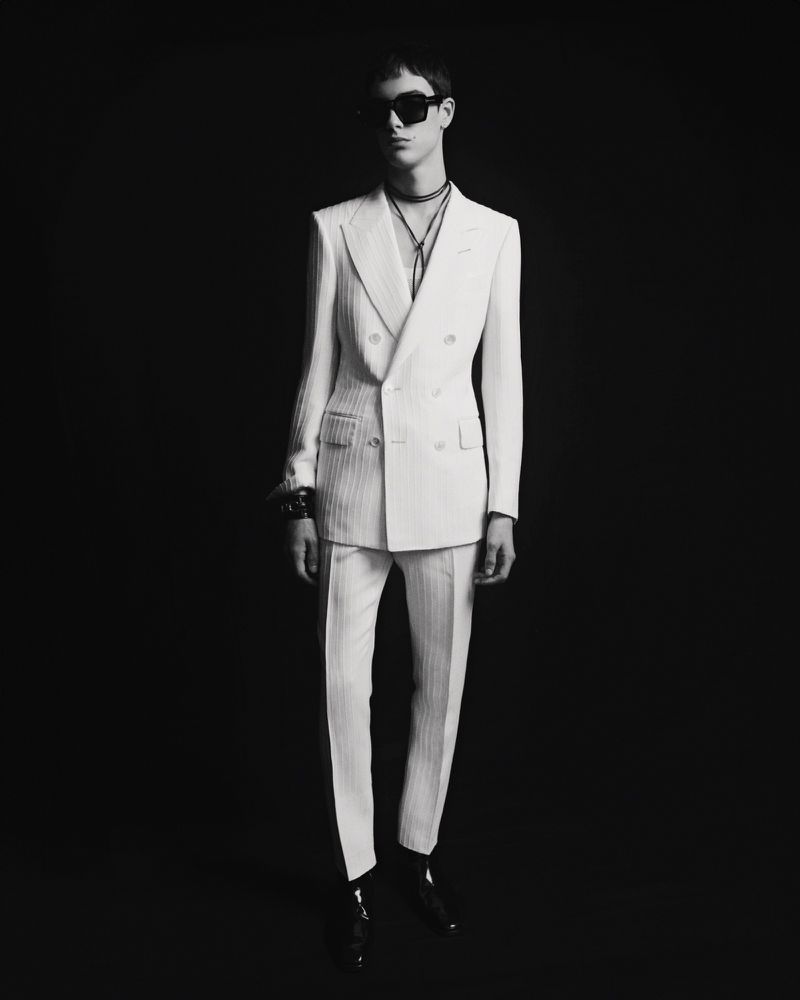 Monochromatic full shot of an adult male model wearing a white double-breasted suit with striped detail, a black necklace, and sunglasses.